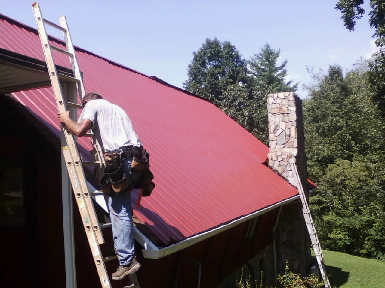 Roofer on a ladder beside a red metal roof and stone chimney — Soffit & Fascia Repair by Roofing Brothers in Streator